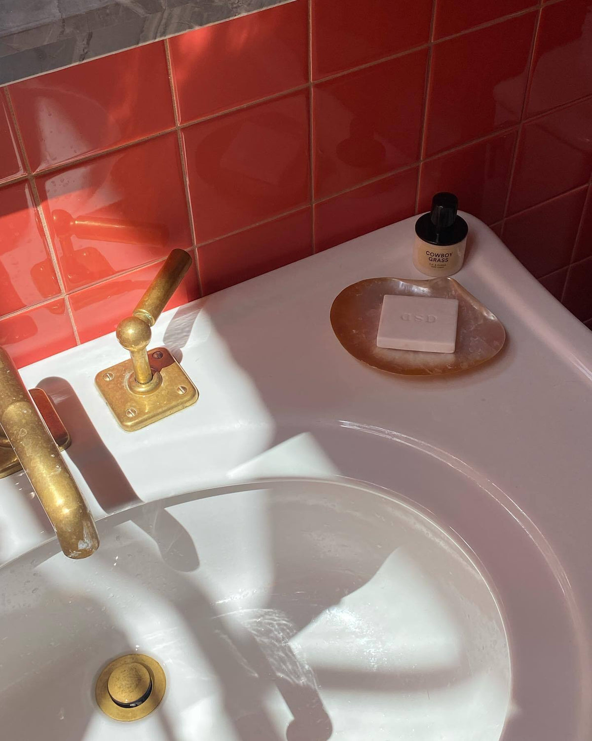 A sunlit pink bathroom sink with brass taps, a bar of soap on a resin dish and a bottle of hand cream, against red tile