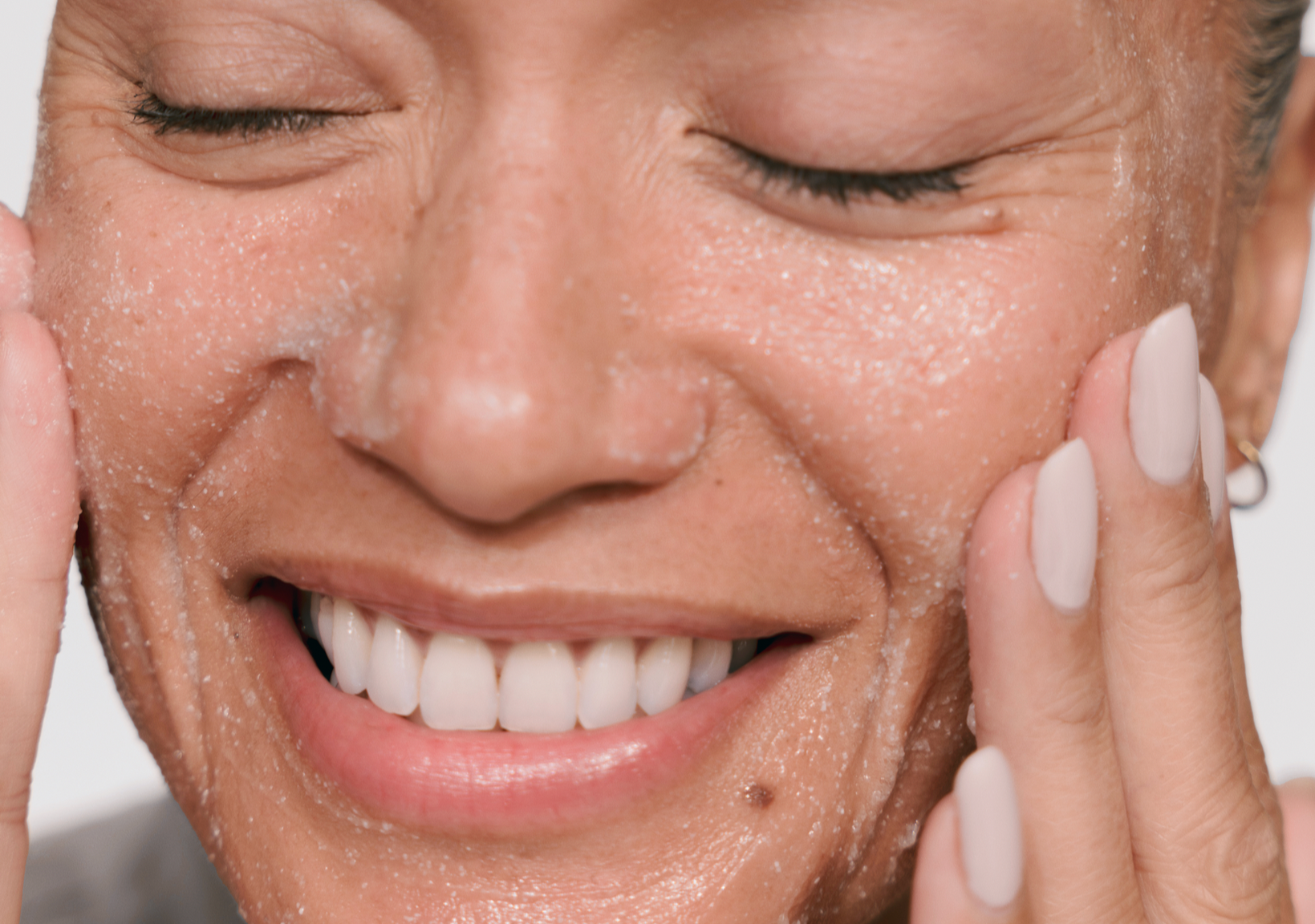 Close-up of a smiling woman massaging an exfoliating scrub onto her cheeks with both hands.