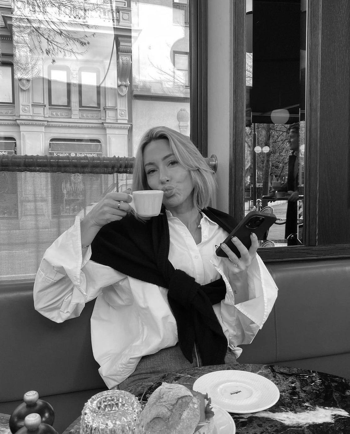 A woman sipping espresso in a cafe window seat, holding her phone