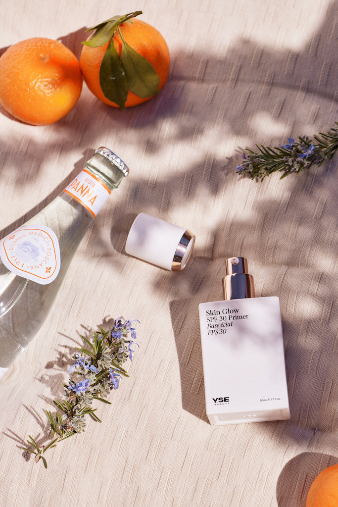 YSE Beauty Skin Glow SPF 30 Primer lying in dappled sunlight beside oranges, rosemary sprigs and a bottled drink