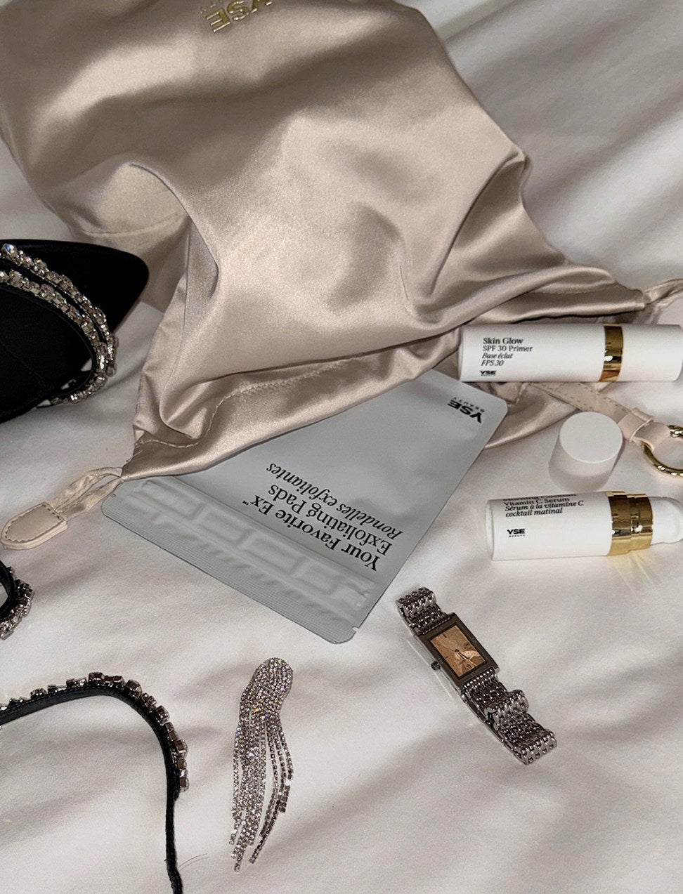 A champagne satin bag of YISE minis on white sheets beside crystal heels and a watch