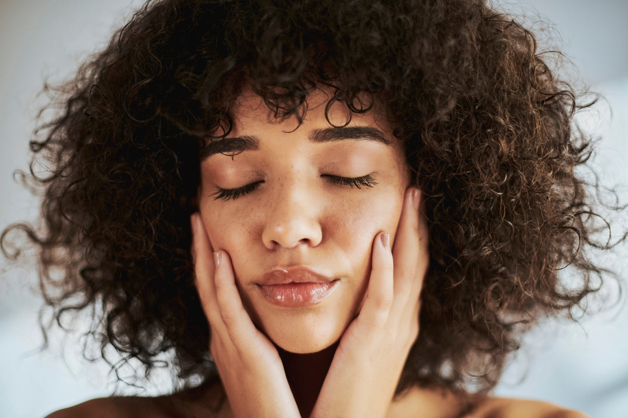 A woman with curly hair, eyes closed, resting both hands against her cheeks